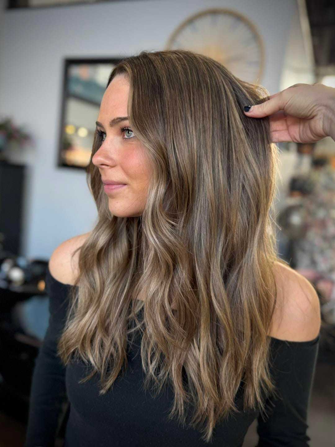 Woman with long wavy brown hair styled in salon, wearing black off-shoulder top.