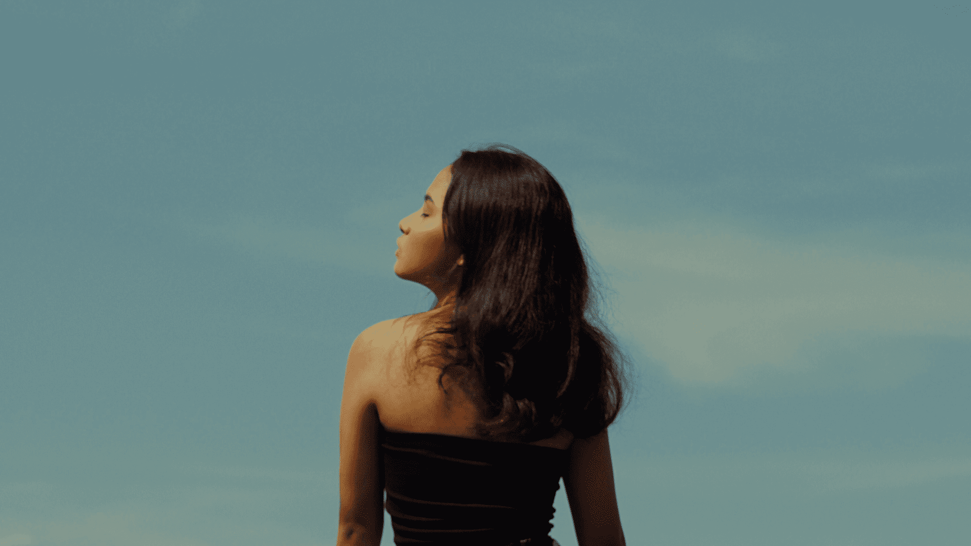 Young woman with long hair gazes at the sky under a blue, cloud-dotted sky.